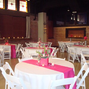 May include: A white tablecloth with a pink runner and white chairs set up for a wedding reception. The room has exposed brick walls and a large window with stained glass.