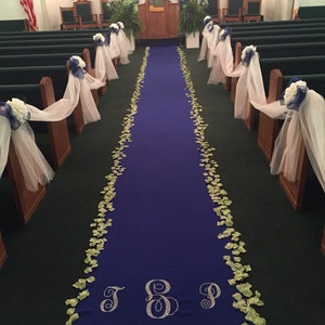 May include: A blue aisle runner with white tulle and floral decorations on the pews. The aisle runner has green flower petals scattered on it and the initials "J & P" and the date "August 8, 2015" are embroidered on it.