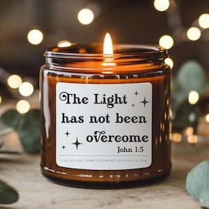 May include: A lit soy wax candle in a brown glass jar. The label reads "The Light has not been overcome" with the Bible verse John 1:5. The candle is 9 oz.