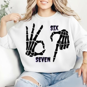 May include: White sweatshirt with a black skeleton hand graphic forming the number six and seven. The word "SIX" is above the hand gesture, and "SEVEN" is below. The graphic has a purple and blue gradient.