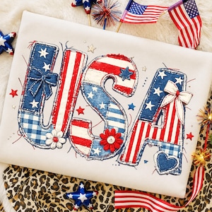 Könnte beinhalten: Ein weißes T-Shirt mit dem Wort "USA" in einem patriotischen Design. Die Buchstaben sind mit rot-weißen Streifen, blauem Denim und weißen Sternen verziert. Das Design umfasst Schleifen, Blumen und ein Herz.