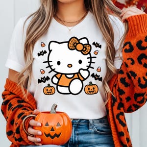 Könnte beinhalten: Ein weißes Kurzarm-T-Shirt mit einer Grafik einer weißen Katzenfigur mit schwarzer Umrandung, die ein orangefarbenes Oberteil und eine Schleife mit Spinnennetz-Muster trägt, umgeben von schwarzen Fledermäusen, Kürbissen und Bonbons.