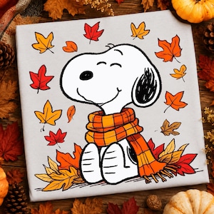 May include: A light gray t-shirt featuring a graphic of a white cartoon dog with a black ear, wearing an orange and yellow plaid scarf. The dog is surrounded by scattered autumn leaves in shades of red, orange, and yellow on the fabric surface.