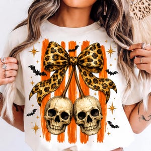 Op de afbeelding: Een crèmekleurig t-shirt met een grafische print van twee menselijke schedels die aan een grote strik met luipaardprint hangen. Het ontwerp bevat verticale oranje penseelstreken, kleine zwarte vleermuizen en gouden sterren op de voorzijde.