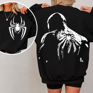 Può includere: Una felpa nera a maniche lunghe con una grande grafica bianca sul retro che raffigura una figura di supereroe stilizzata con un emblema a ragno. La parte anteriore della felpa presenta un logo a ragno bianco più piccolo sul petto.