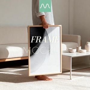 May include: A rectangular wooden frame mockup with the text "FRAME MOCKUP" in white. The artwork inside the frame features a black and white abstract design. The frame is held in front of a person in a light-colored room with a sofa and side table.