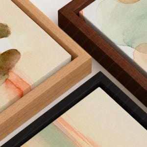 May include: Three rectangular canvas prints featuring abstract watercolor designs in shades of sage green, soft orange, and cream. Each canvas is encased in a distinct floating frame, with one light wood, one dark wood, and one black frame finish.