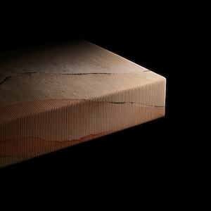 Puede incluir: Un bloque rectangular con una superficie texturizada y acanalada con l&iacute;neas horizontales. El objeto muestra un degradado de tonos beige, marr&oacute;n claro y naranja terracota con finas grietas horizontales oscuras e irregulares.