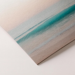 May include: A print of a beach scene with a turquoise wave breaking on the shore. The sky is a soft pink and the sand is a light brown. The print has a textured surface.