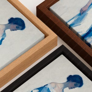 May include: Three rectangular canvas prints featuring blue-toned figurative artwork, each encased in a different floating frame: one light wood, one dark brown wood, and one black frame. The artwork depicts a person with dark hair in profile.