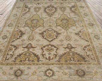 Könnte beinhalten: Ein großer rechteckiger Teppich mit einem komplizierten, symmetrischen Muster. Der Teppich hat einen beigen Hintergrund mit braunen und goldenen floralen und geometrischen Designs. Der Rand ist ein dunklerer Beigeton mit ähnlichen Details. Der Teppich liegt auf einem Holzboden.