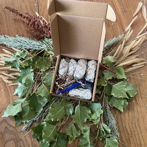 May include: A small, open cardboard box containing several foil-wrapped items and a blue object, nestled on shredded paper. The box is surrounded by green leaves, dried grasses, and other natural elements, arranged on a wooden surface.