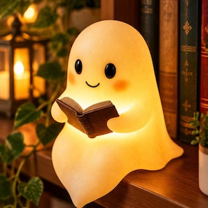May include: A glowing, translucent, off-white ghost-shaped lamp with a smooth surface, black oval eyes, a small curved mouth, and pink cheeks. The ghost figure is holding a small, dark brown open book in its hands, emitting a warm light.