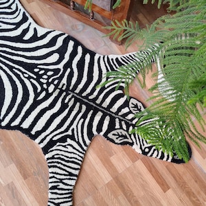 May include: A black and white zebra-print rug, shaped like a zebra, lies on a wooden floor. The rug's design mimics a zebra's stripes. Green foliage is in the upper right corner. The rug is a decorative item for a home.