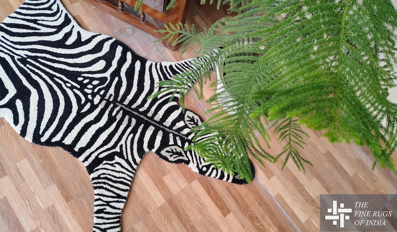 May include: A black and white zebra-print rug, shaped like a zebra, lies on a wooden floor. The rug's design mimics a zebra's stripes. Green foliage is in the upper right corner. The rug is a decorative item for a home.
