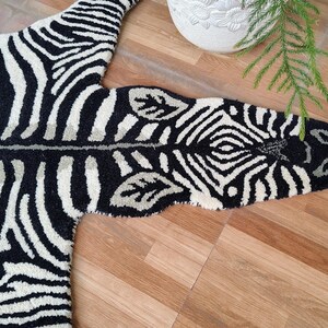May include: A black and white zebra-striped rug with a unique shape, resembling a zebra hide. The rug is made of a soft material and is placed on a wooden floor. The Fine Rugs of India logo is in the bottom right corner.