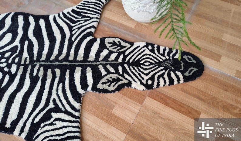 May include: A black and white zebra-striped rug with a unique shape, resembling a zebra hide. The rug is made of a soft material and is placed on a wooden floor. The Fine Rugs of India logo is in the bottom right corner.