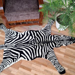 May include: A black and white zebra-patterned rug with a detailed head and legs. The rug is placed on a wooden floor, with plants and a chair in the background. The rug is a decorative item.