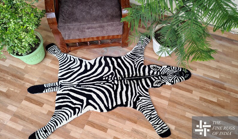 May include: A black and white zebra-patterned rug with a detailed head and legs. The rug is placed on a wooden floor, with plants and a chair in the background. The rug is a decorative item.