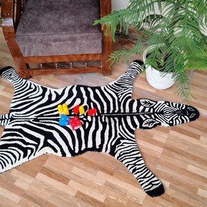 May include: A black and white zebra-patterned rug with a zipper down the middle. The rug is shaped like a zebra with legs and a head. Colorful toy blocks are placed on the rug. The rug is on a wooden floor.