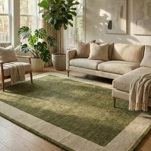 May include: A living room with a green rug with a beige border. The room features a beige sectional sofa, a wooden armchair, and potted plants. Large windows with beige curtains provide natural light.