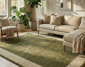 May include: A rectangular area rug featuring a deep olive green center panel framed by a wide, solid beige border. The rug is positioned on a light-toned wooden floor in a room with a beige sofa, a matching armchair, and two large potted plants.