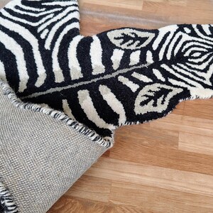 May include: A black and white zebra-striped rug with a leaf-shaped design. The rug is made of a thick, textured material and has a gray backing. The rug is partially rolled up, revealing the underside.