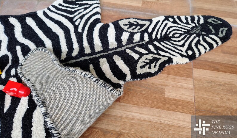 May include: A black and white zebra-striped rug with a leaf-shaped design. The rug is made of a thick, textured material and has a gray backing. The rug is partially rolled up, revealing the underside.