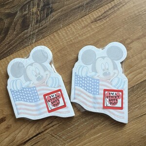 May include: Two die-cut sticky note pads shaped like Mickey Mouse holding an American flag. Each pad features a red square sticker with the text I'M A STICKY NOTE PAD! and a small graphic of a page corner being peeled back, with Disney branding.
