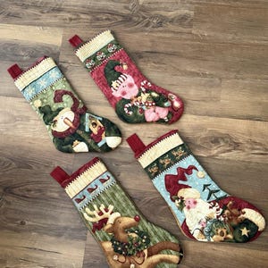 Puede incluir: Cuatro calcetines navideños acolchados con diseños festivos. Uno muestra un muñeco de nieve, otro un cerdo con bastones de caramelo, uno un reno con una corona y el último una figura de Santa Claus. Cada calcetín tiene un lazo rojo para colgar.
