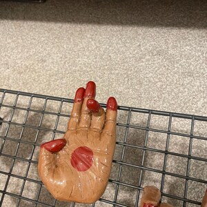 May include: A ceramic hand sculpture with a brown skin tone and red fingernails. A red circle is painted on the palm. The hand is resting on a black wire rack.