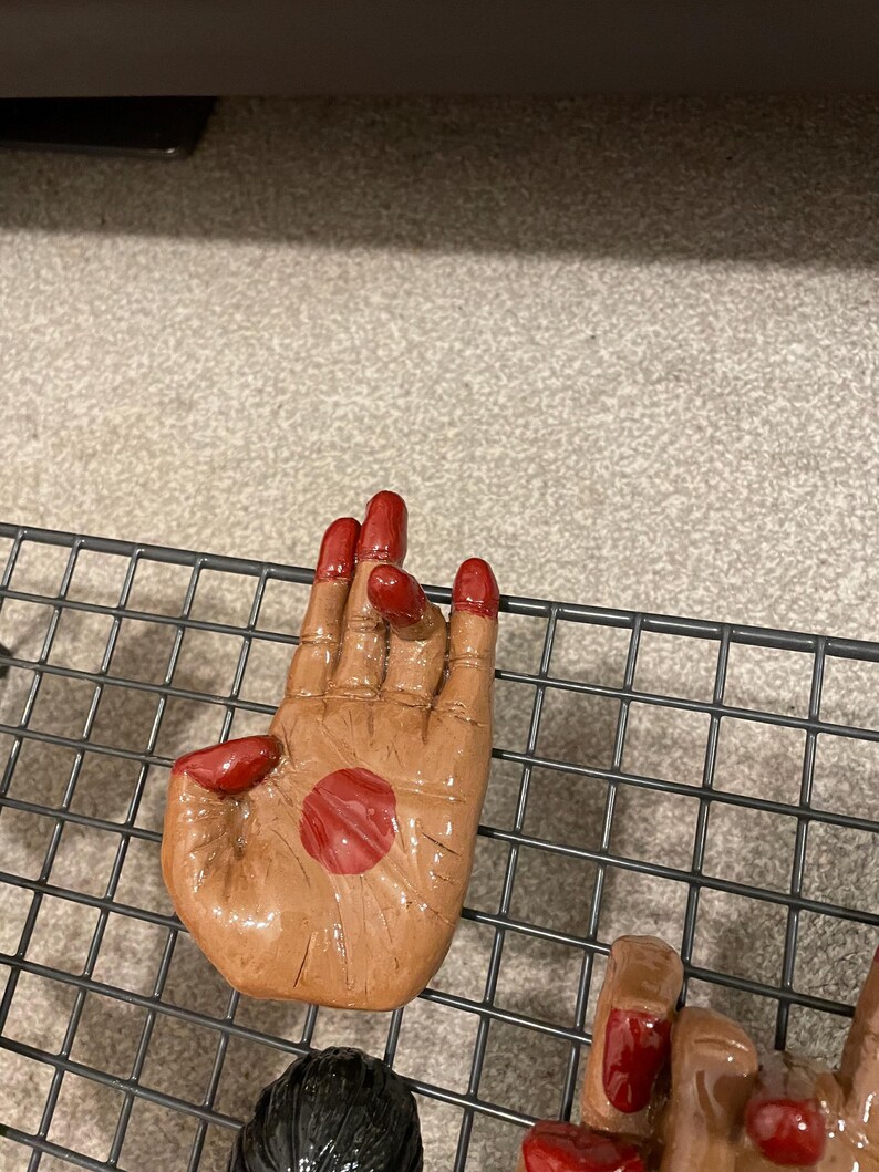 May include: A ceramic hand sculpture with a brown skin tone and red fingernails. A red circle is painted on the palm. The hand is resting on a black wire rack.