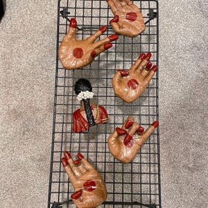 May include: A black metal grid displaying five hand-shaped decorative pieces and a small bust. The hands are tan with red nail polish and a red dot on the palm. The bust is red and black with white flowers.