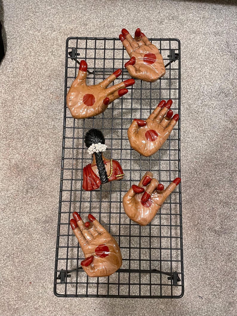 May include: A black metal grid displaying five hand-shaped decorative pieces and a small bust. The hands are tan with red nail polish and a red dot on the palm. The bust is red and black with white flowers.