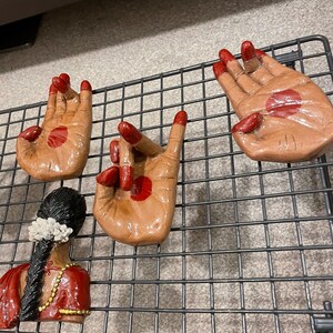 May include: Four brown ceramic hand sculptures with red accents, arranged on a black metal grid. The hands are in various mudra positions. A sculpture of a woman in a red sari is also present.