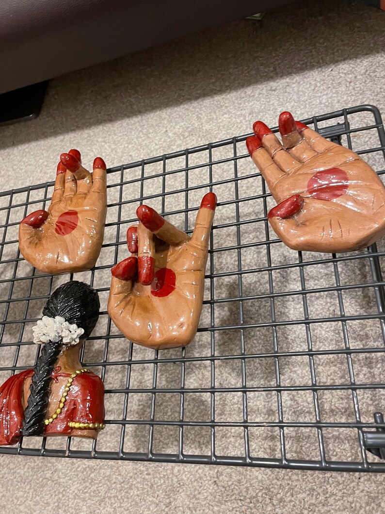 May include: Four brown ceramic hand sculptures with red accents, arranged on a black metal grid. The hands are in various mudra positions. A sculpture of a woman in a red sari is also present.