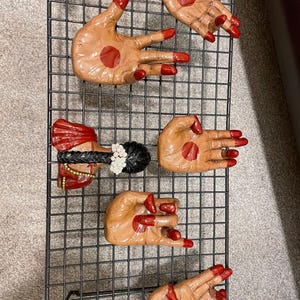 May include: A collection of hand-shaped decorative items with red nail polish and a red dot on the palm. One item depicts a woman's head with a red and gold headdress and black hair. The items are arranged on a black wire grid.