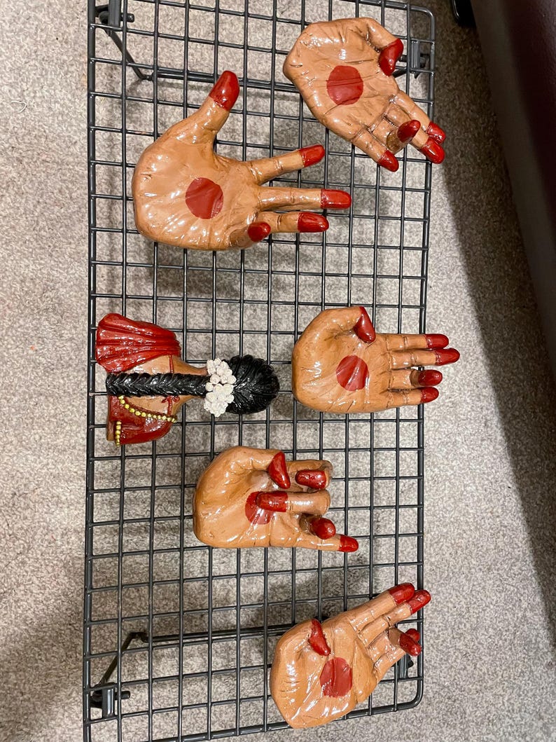 May include: A collection of hand-shaped decorative items with red nail polish and a red dot on the palm. One item depicts a woman's head with a red and gold headdress and black hair. The items are arranged on a black wire grid.