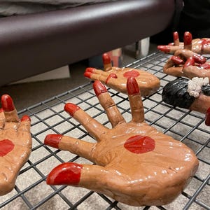 May include: Hand-shaped ceramic sculptures with brown skin tones, red fingertips, and a red dot on the palm. The hands are arranged on a metal rack, showcasing the details of the artwork.