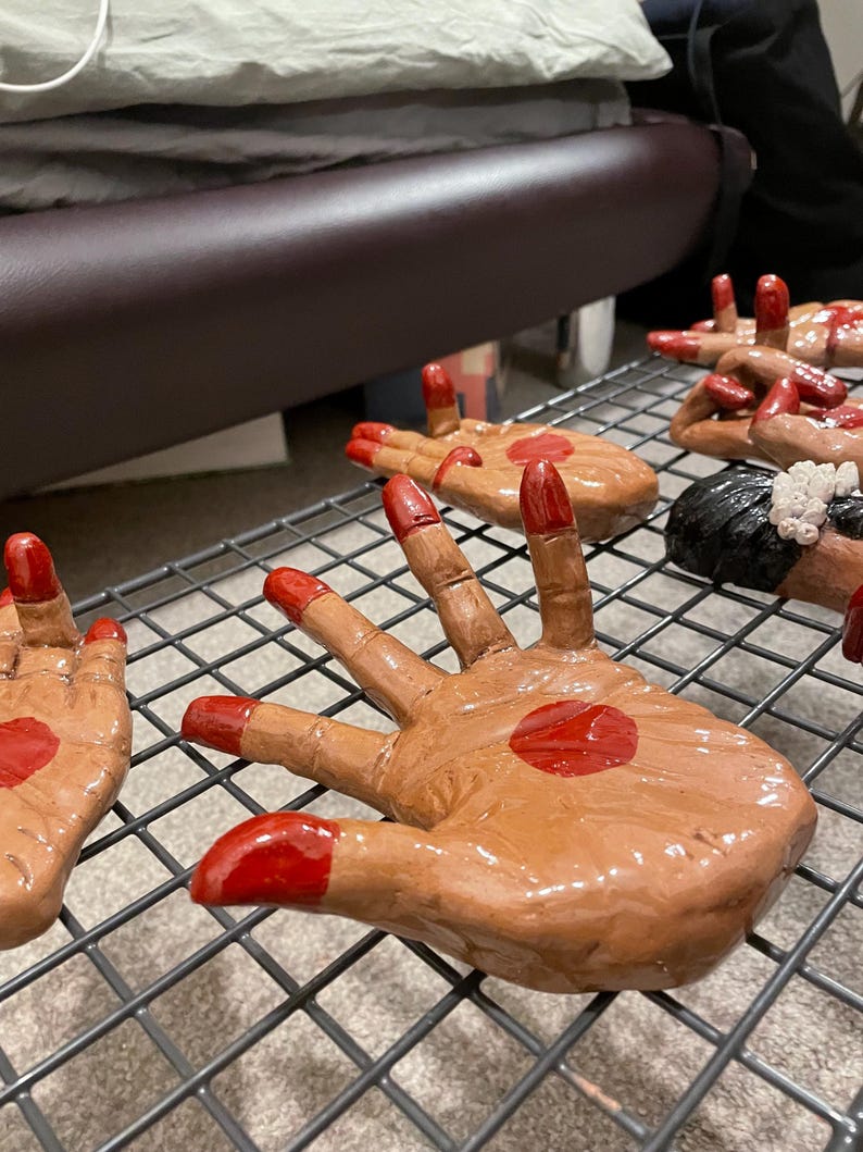 May include: Hand-shaped ceramic sculptures with brown skin tones, red fingertips, and a red dot on the palm. The hands are arranged on a metal rack, showcasing the details of the artwork.