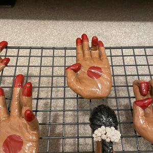 May include: Hand-shaped ceramic figurines with red nail polish and a red dot on the palm. The figurines are brown and are arranged on a black wire rack. The image also shows a woman with black hair and a red sari.