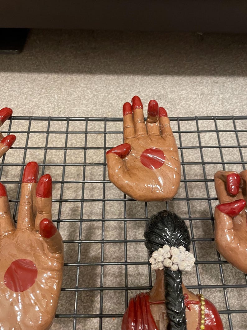 May include: Hand-shaped ceramic figurines with red nail polish and a red dot on the palm. The figurines are brown and are arranged on a black wire rack. The image also shows a woman with black hair and a red sari.