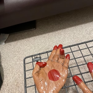 May include: A brown ceramic hand sculpture with red painted fingernails and a red dot on the palm. The sculpture is placed on a metal wire rack.