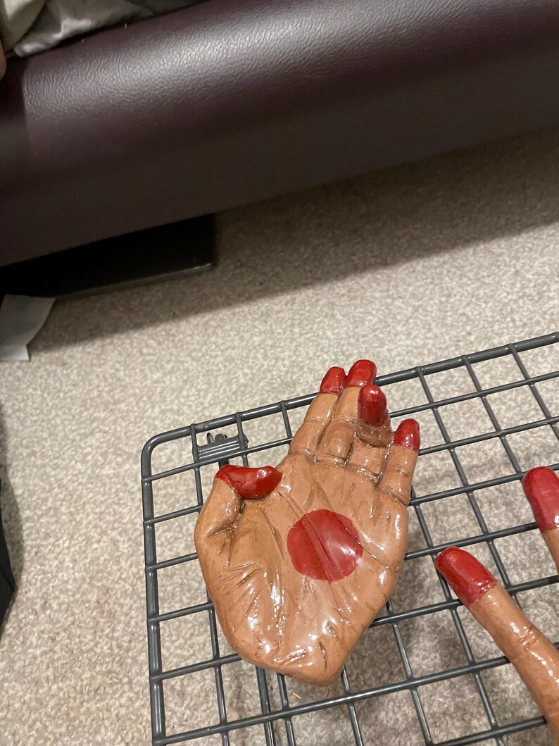May include: A brown ceramic hand sculpture with red painted fingernails and a red dot on the palm. The sculpture is placed on a metal wire rack.