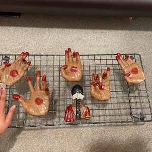 May include: A collection of brown hand-shaped figurines with red accents on a black wire rack. The figurines have red painted fingernails and a red dot on the palm. A small figurine of a woman in a red sari is also present.