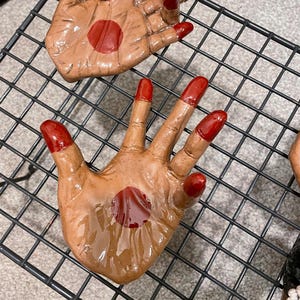 May include: Hand-shaped ceramic art pieces with a brown skin tone and bright red accents. The fingertips and a central palm circle are painted red. The pieces are arranged on a black wire rack.