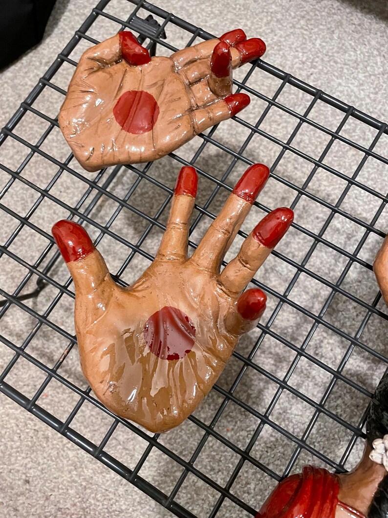 May include: Hand-shaped ceramic art pieces with a brown skin tone and bright red accents. The fingertips and a central palm circle are painted red. The pieces are arranged on a black wire rack.