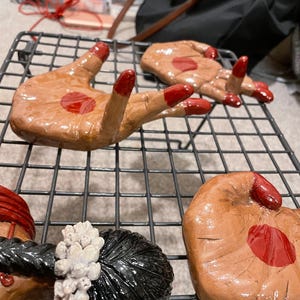 May include: Hand-shaped ceramic sculptures with brown skin tones and red accents. The fingernails and a central palm detail are painted red. The sculptures are arranged on a black wire rack.