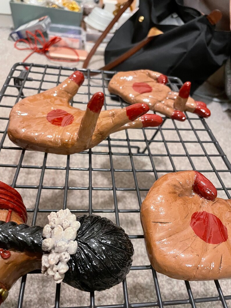 May include: Hand-shaped ceramic sculptures with brown skin tones and red accents. The fingernails and a central palm detail are painted red. The sculptures are arranged on a black wire rack.