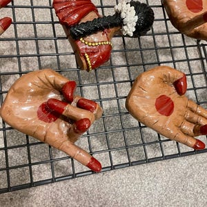 May include: Hand-shaped ceramic figurines with brown skin tones, red nail polish, and red dot accents. Some figurines have a red head covering and gold jewelry. The figurines are arranged on a black wire rack.